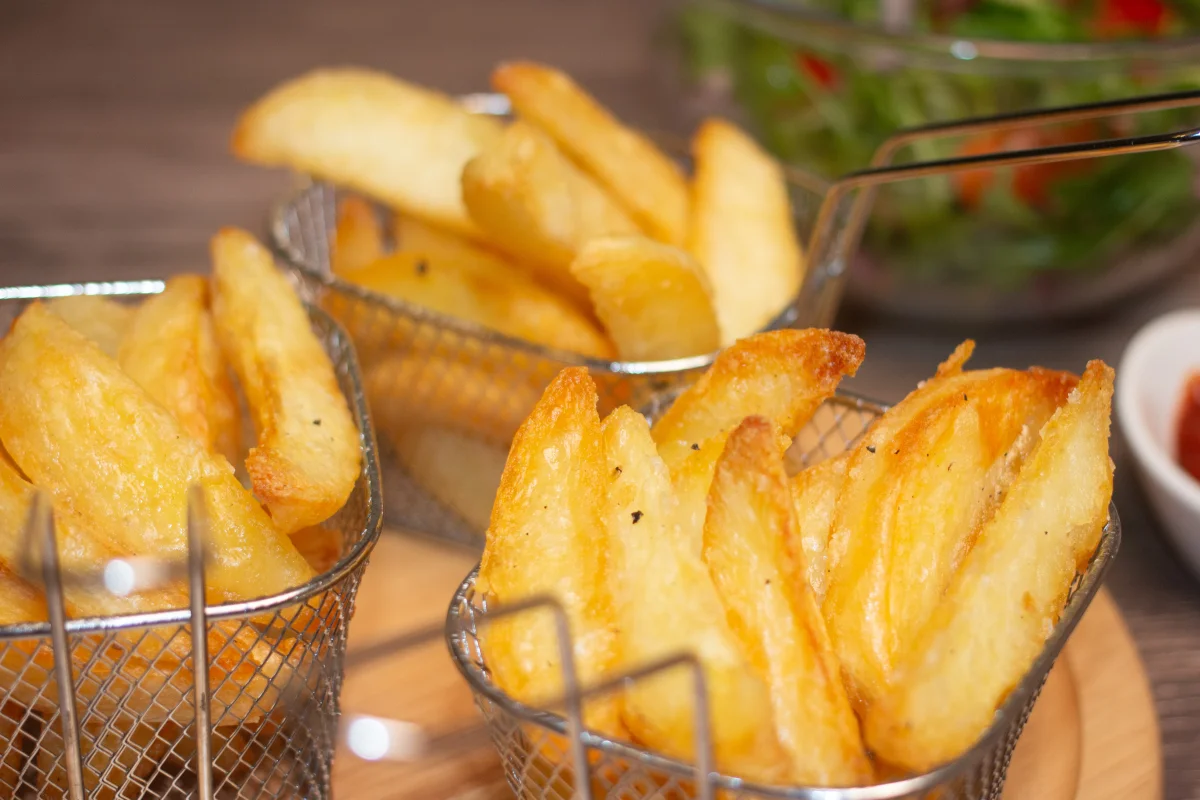 Three portions of wedges in small metal baskets on a circular board, a bowl of salad and a bbq sauce sauce are blurred in the background.
